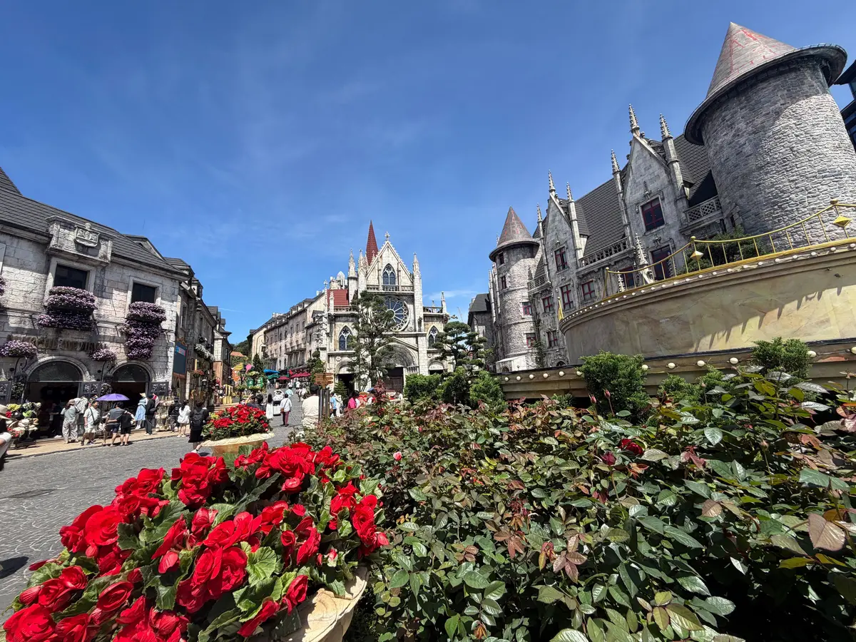 赤やピンクのバラが茂る花壇の先に、中世ヨーロッパ風の建物が並ぶ風景。青空を背景に、塔やアーチ型の装飾が施された建物が特徴的。人々が散策している。ベトナム・ダナンの人気観光地「フランス村」の写真。