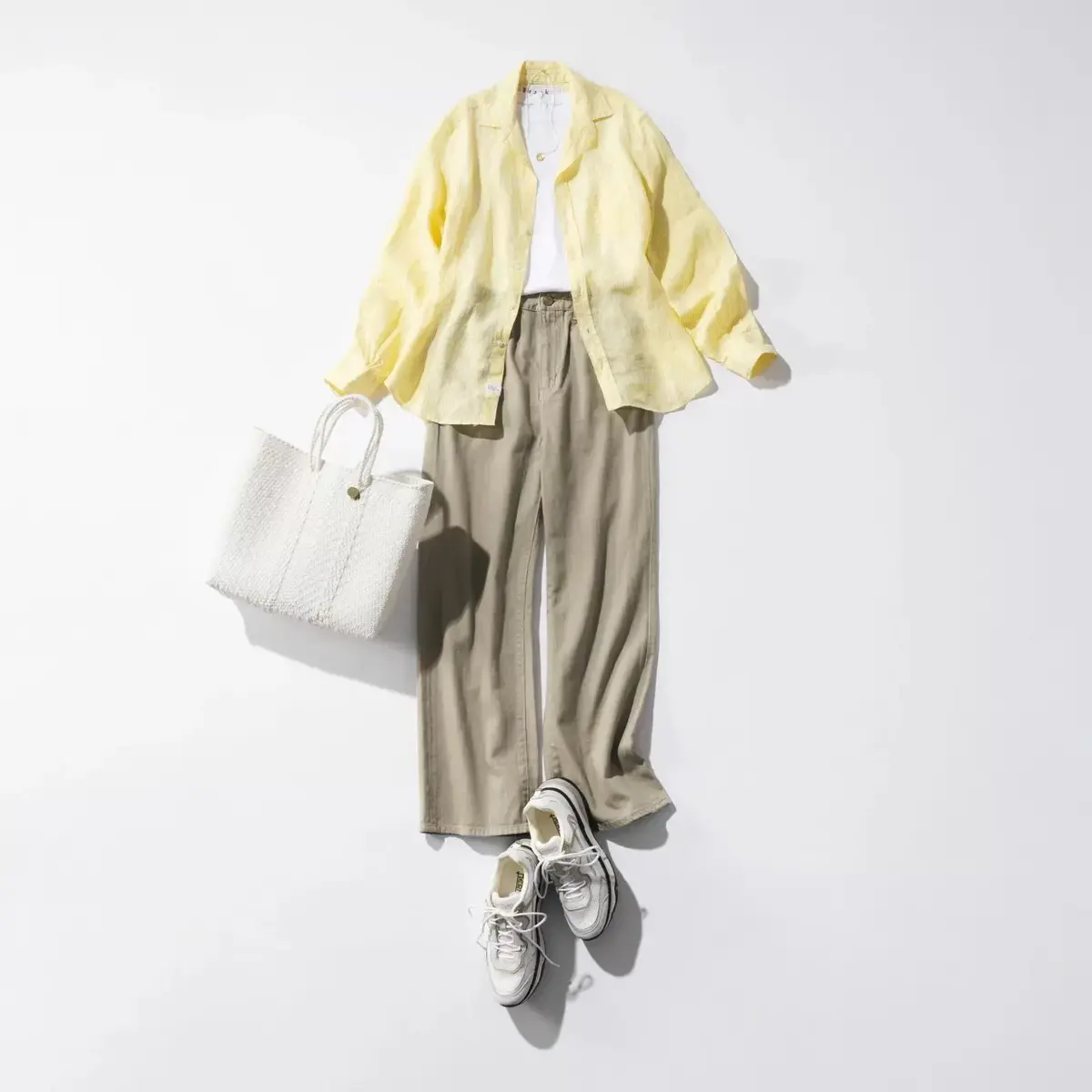 黄色のリネンシャツ、ベージュのワイドパンツ、白いTシャツ、白いスニーカー、白いかごバッグが平らな白い背景に置かれている。全体的に淡い色合いで、春らしいコーディネート。