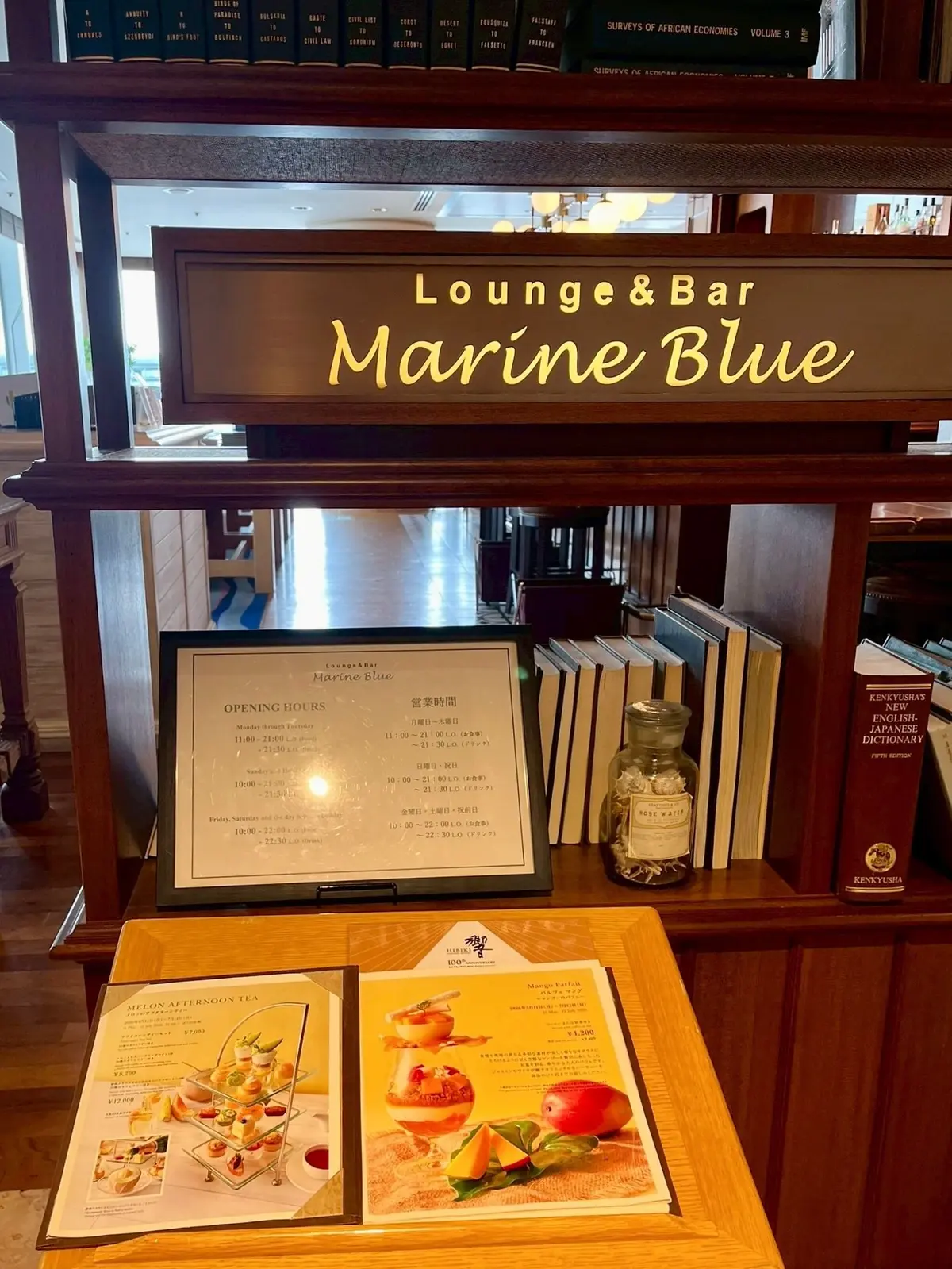 ラウンジ＆バーの看板とメニューが置かれた木製の棚、背景には本が並ぶ