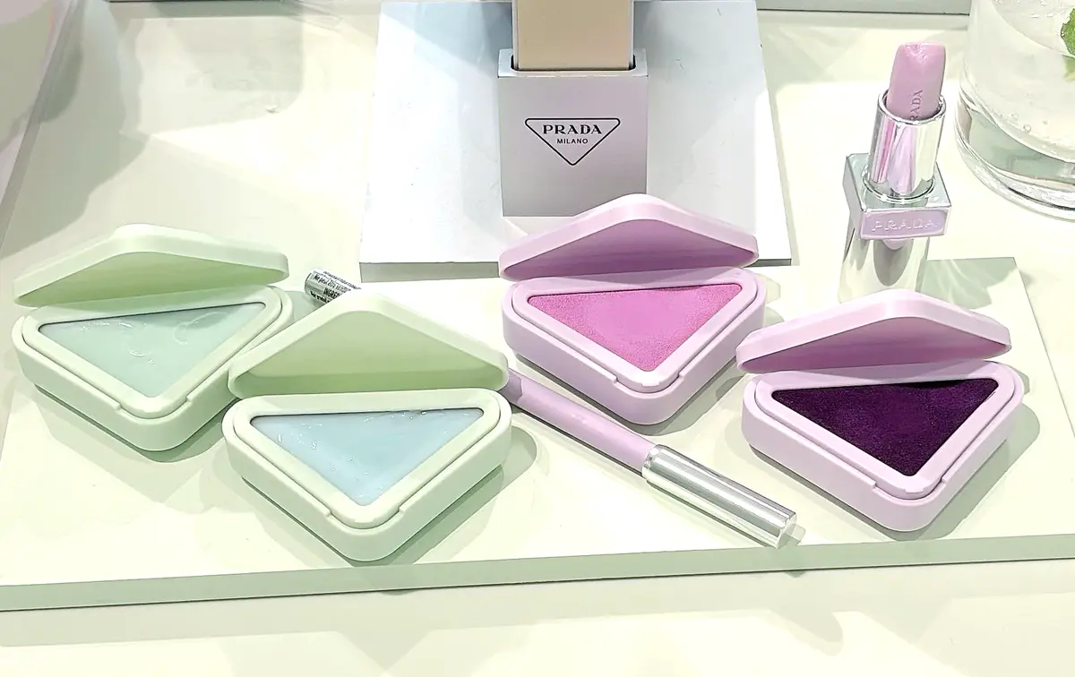 PRADAの化粧品。三角形のパウダーケースが緑、ピンク、紫の3色。中央にはPRADAのロゴが入った白い箱と、ピンク色のリップスティックが置かれている。