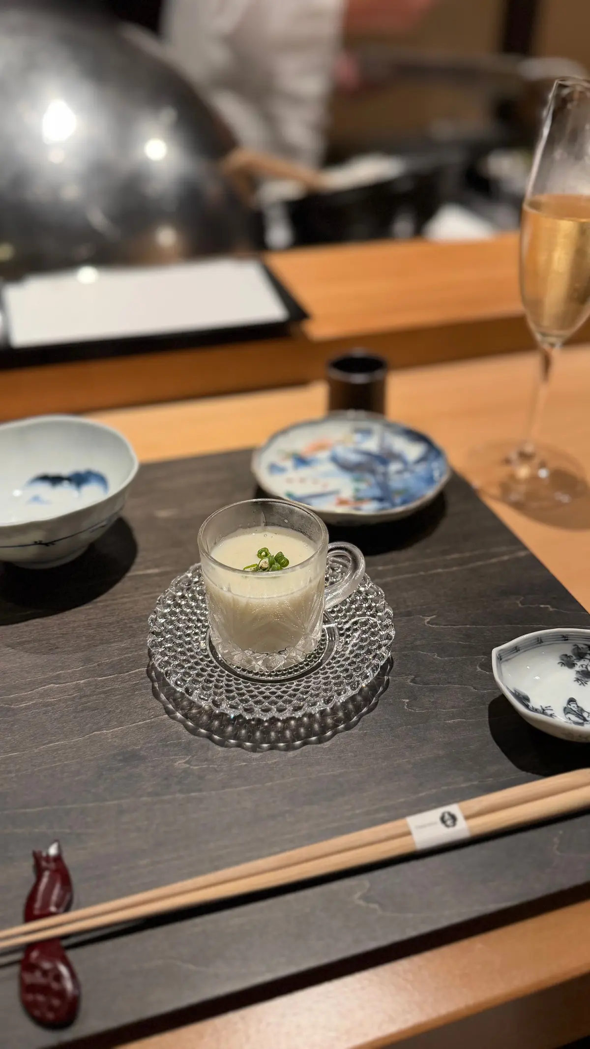 木製のテーブルに置かれた、透明なカップに入った白いスープ。緑色の薬草が浮かび、銀色のソーサーに乗っている。周辺には小皿やグラス、箸が配置されている。