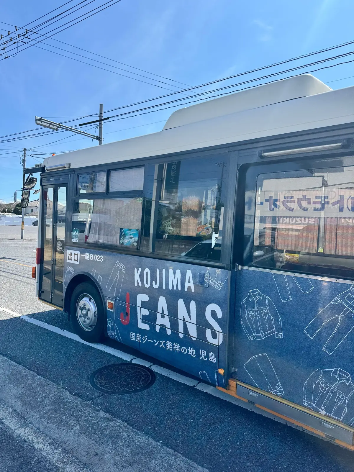 青空の下、白い屋根を持つバスの側面。バスには「KOJIMA JEANS」の文字と、ジーンズのイラストが描かれている。道路脇に停車し、マンホールが写っている。