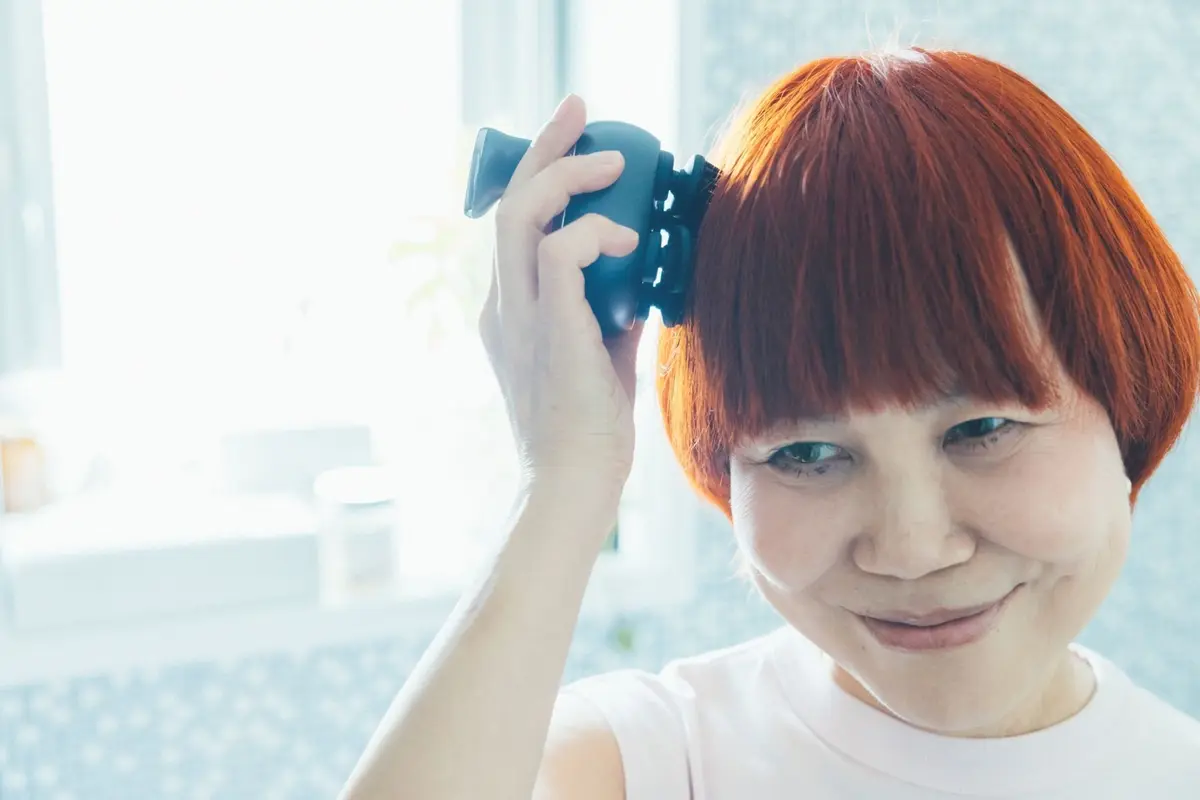 赤毛の女性が、黒い双眼鏡を頭に乗せて微笑んでいる。白いTシャツを着ており、背景には窓と壁がある。