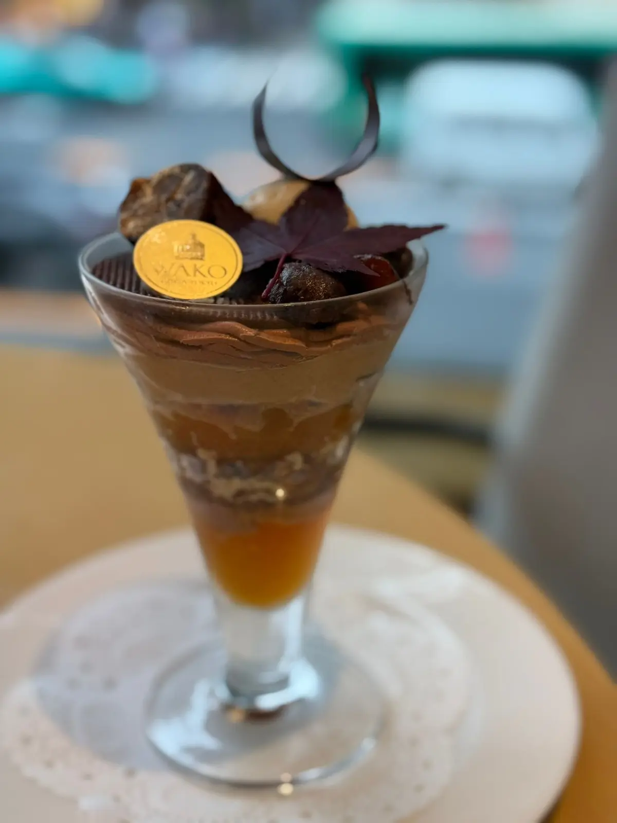 透明なグラスに盛り付けられた、チョコレートとカラメル色の層状デザート。上部にはチョコレートの飾りと、赤紫色の葉、ワコールのロゴが入った丸いシールが飾られている。白い模様のソーサーに乗せられている。