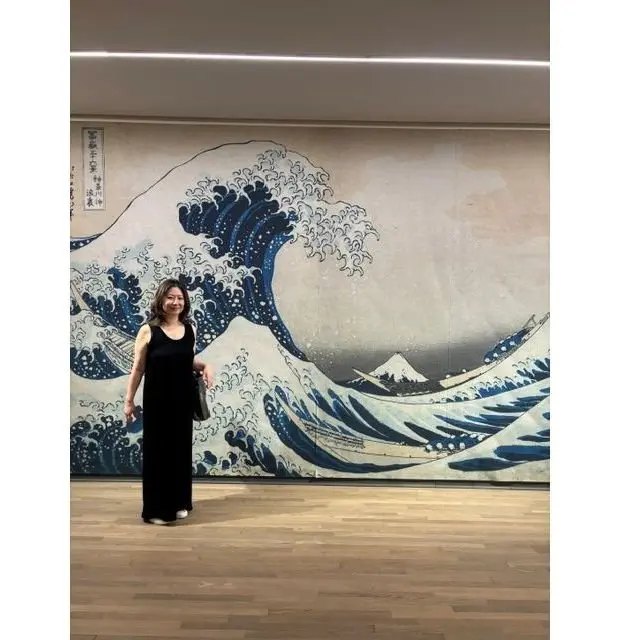 美術館で、黒いワンピースを着た女性が、葛飾北斎の「神奈川沖浪裏」の前に立って微笑んでいる。波の大きな絵画の前に、女性が正面を向いている。