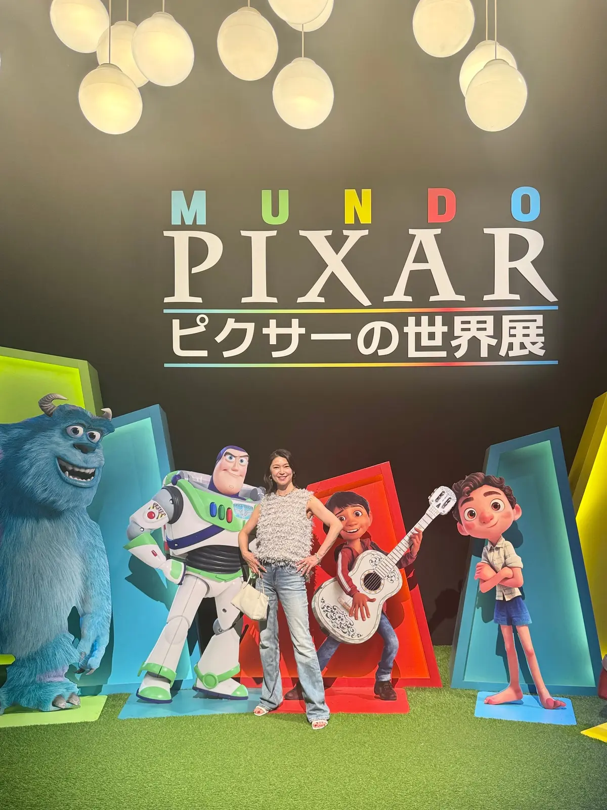 ピクサーの世界「MUNDO PIXAR」の展示で、サリー、バズ・ライトイヤー、ギターを弾く女性、そして映画『Luca』のキャラクターが並んで写っている。背景には球体の照明が吊り下げられている。