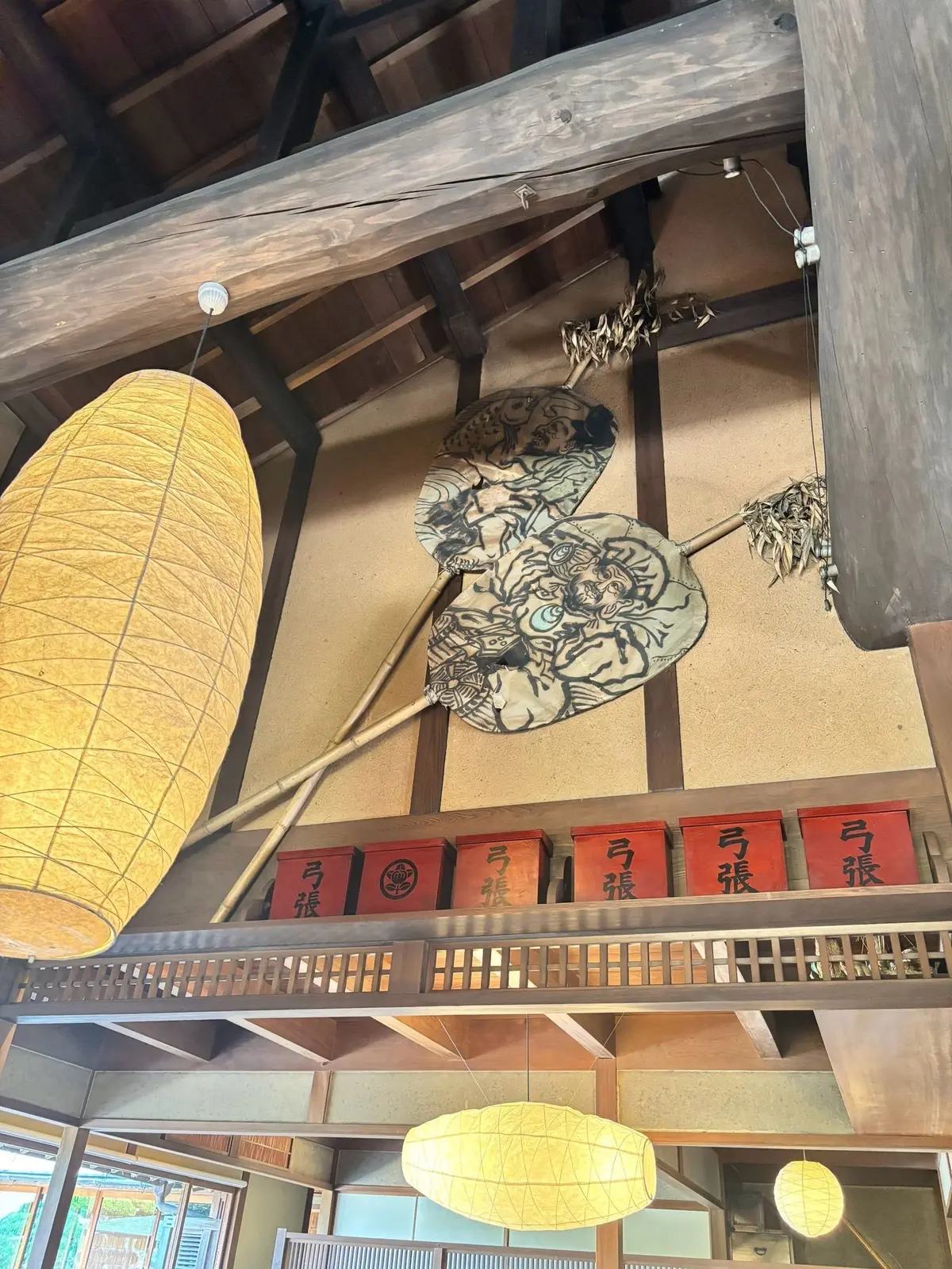 木造の柱と梁が見える屋内。天井には大きな灯籠、壁には白黒の鬼の絵が飾られ、下には赤い提灯が並ぶ。手すりの上には球状の照明が吊り下げられている。