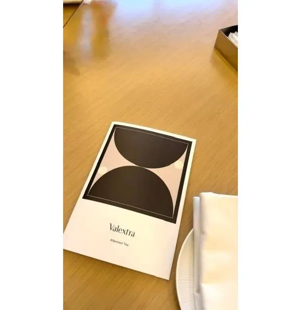木製のテーブルに置かれた、Valextra（ヴァレクストラ）のアフタヌーンティーのメニュー。黒と白の幾何学模様がデザインされており、下部にブランド名が記載されている。ナプキンとソーサーも写っている。