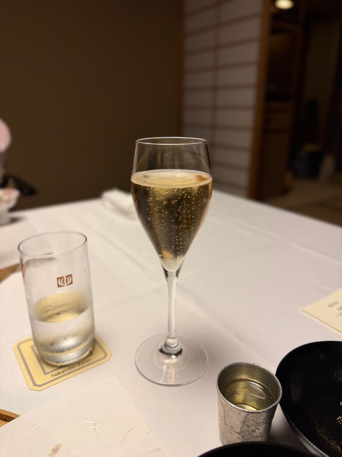 白テーブルに置かれた、シャンパングラスとグラス、小皿。シャンパングラスには泡が立ち、グラスには水が注がれている。背景には木製の障子が見える。
