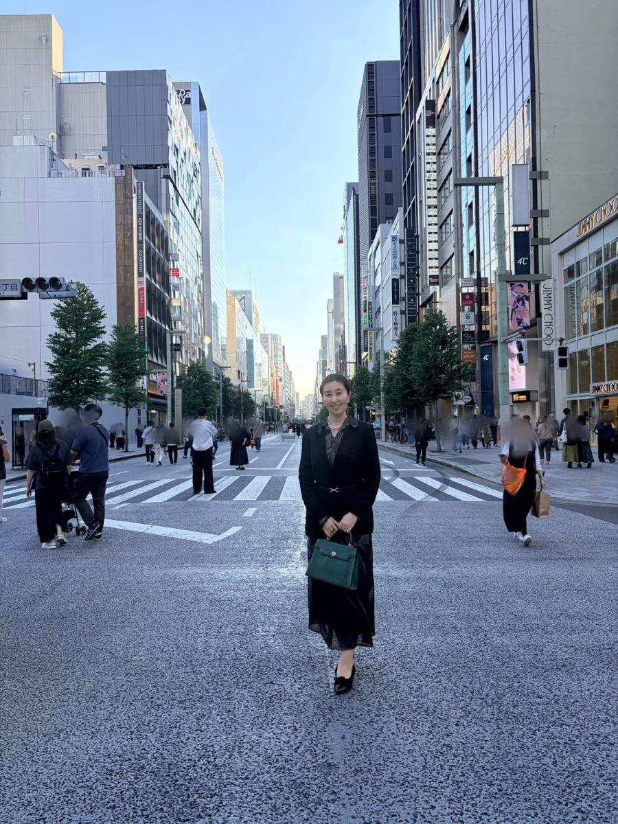 黒いジャケットとロングスカートの女性が、横断歩道を歩いている。背景には高層ビルが立ち並ぶ都市の風景。右手に緑色のハンドバッグを持っている。