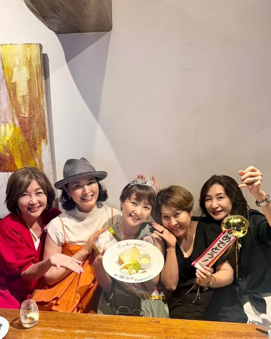 5人の女性がレストランで誕生日ケーキを囲んで笑顔。中央の女性はティアラをつけ、ケーキには日本語でメッセージが書かれている。背景には木製の柱が見える。