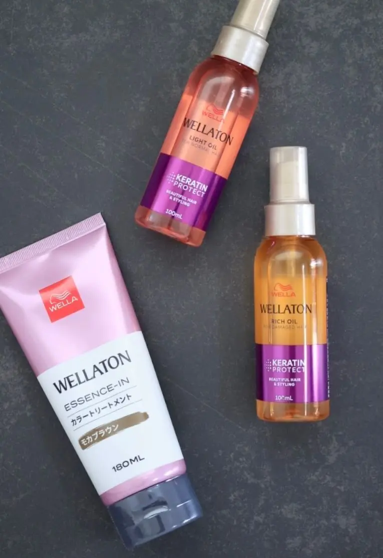 グレーの背景に置かれた、ウェルアトン（Wellaton）のヘアケア製品3点。左はピンクと白のチューブ容器に入った「エッセンス・イン」、右上のオレンジ色のボトルは「ライトオイル」、右下は濃いオレンジ色のボトルで「リッチオイル」。