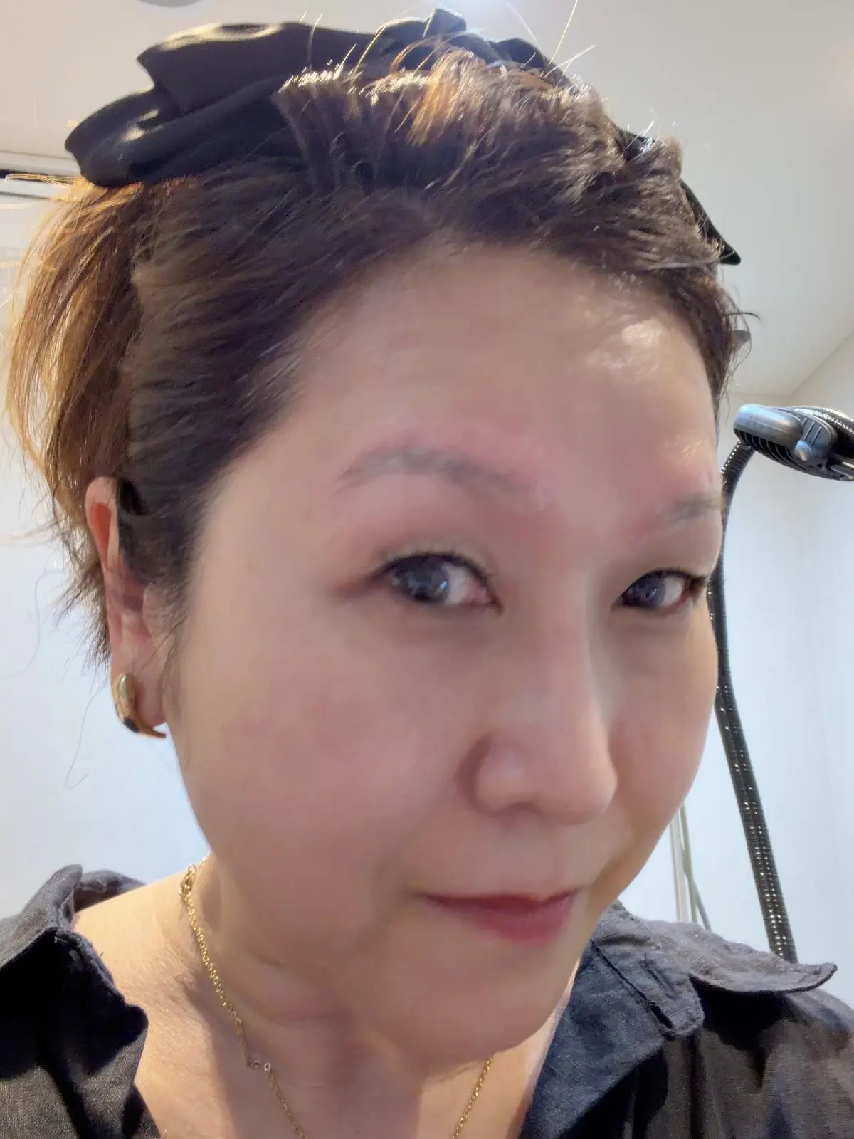 茶髪の女性が正面を向いて写っているセルフィー。眉は自然な茶色で、やや太め。金色のネックレスを着用し、黒いヘアバンドをしている。背景は白っぽい壁。