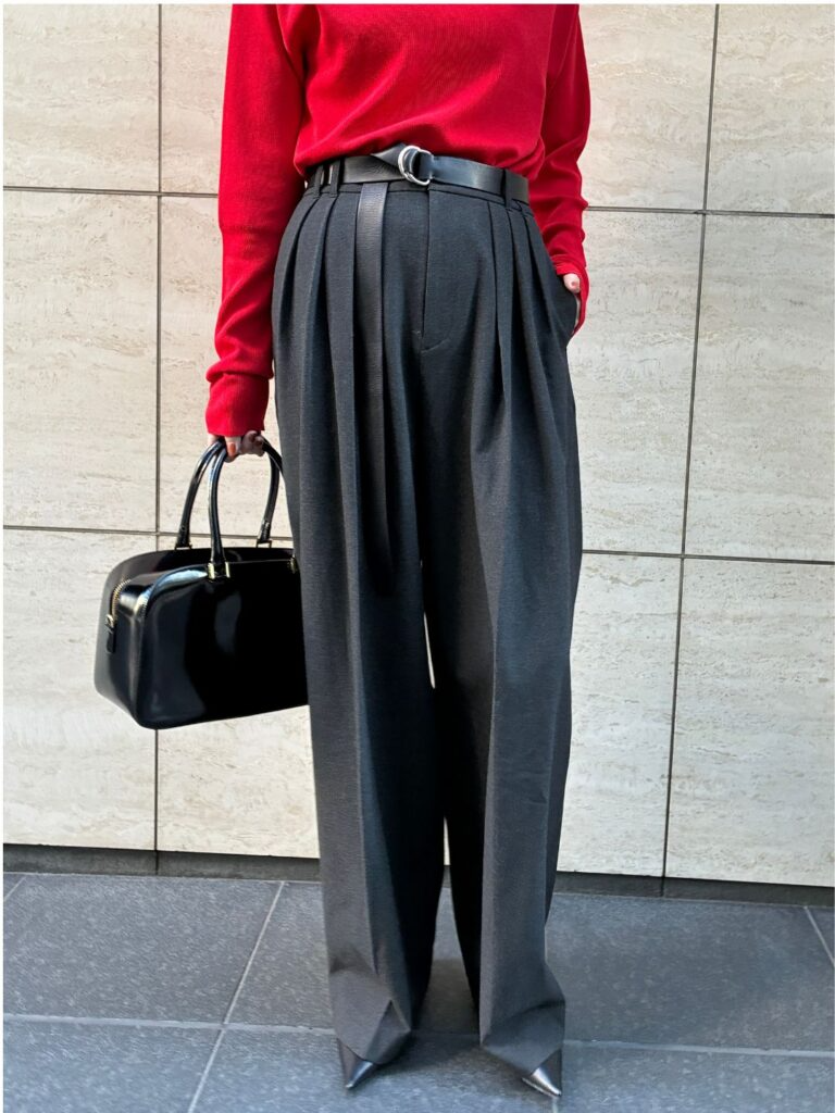 赤いニットにグレーのプリーツパンツを合わせたコーディネート。黒のベルトがウエストマークされており、黒いボストンバッグを持つ。背景はベージュのタイル壁。