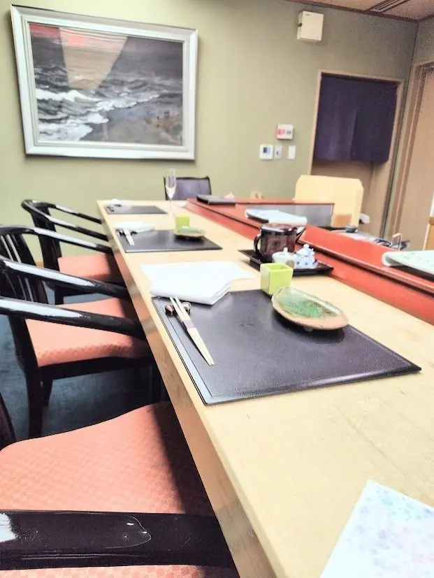 和室の長テーブルに料理が並び、椅子が配置されている。テーブルには緑の小皿、茶碗、食器が置かれ、壁には水彩画が飾られている。