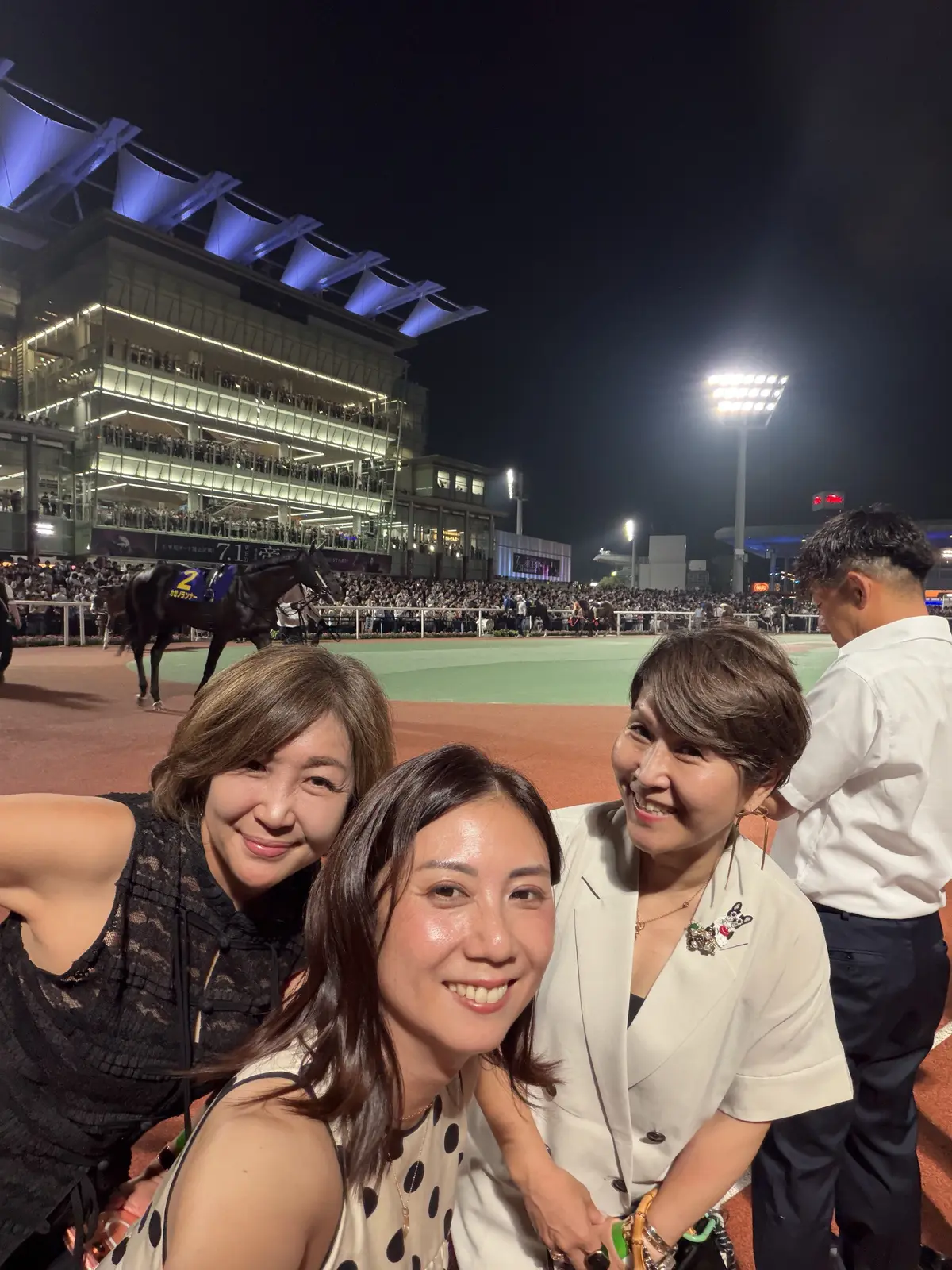 夜の競馬場で、3人の女性が笑顔でセルフィーを撮っている。背景には馬が走り、観客席と照明が広がる。