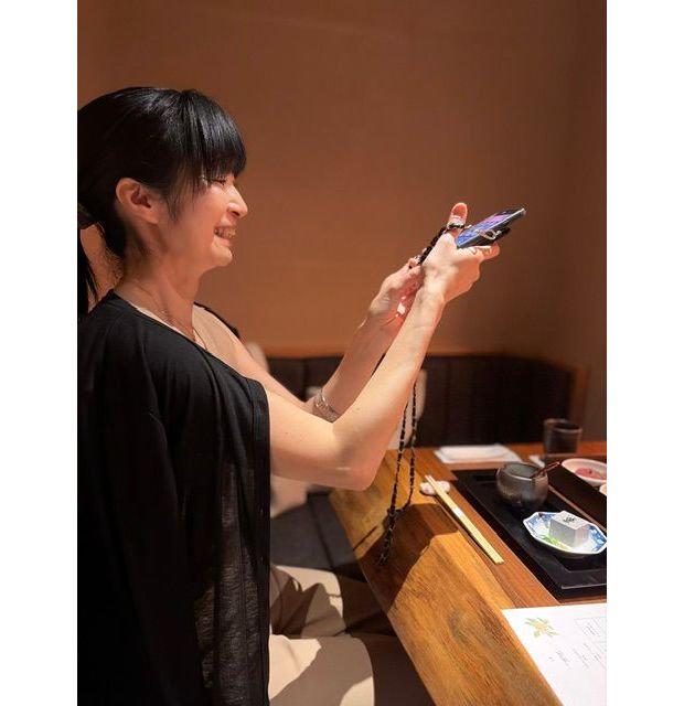 黒いトップスを着た女性が、テーブルに置かれた料理をスマートフォンで撮影している。女性は笑顔で、右手にスマートフォンを持っている。テーブルには箸や料理が並び、背景はベージュ色。