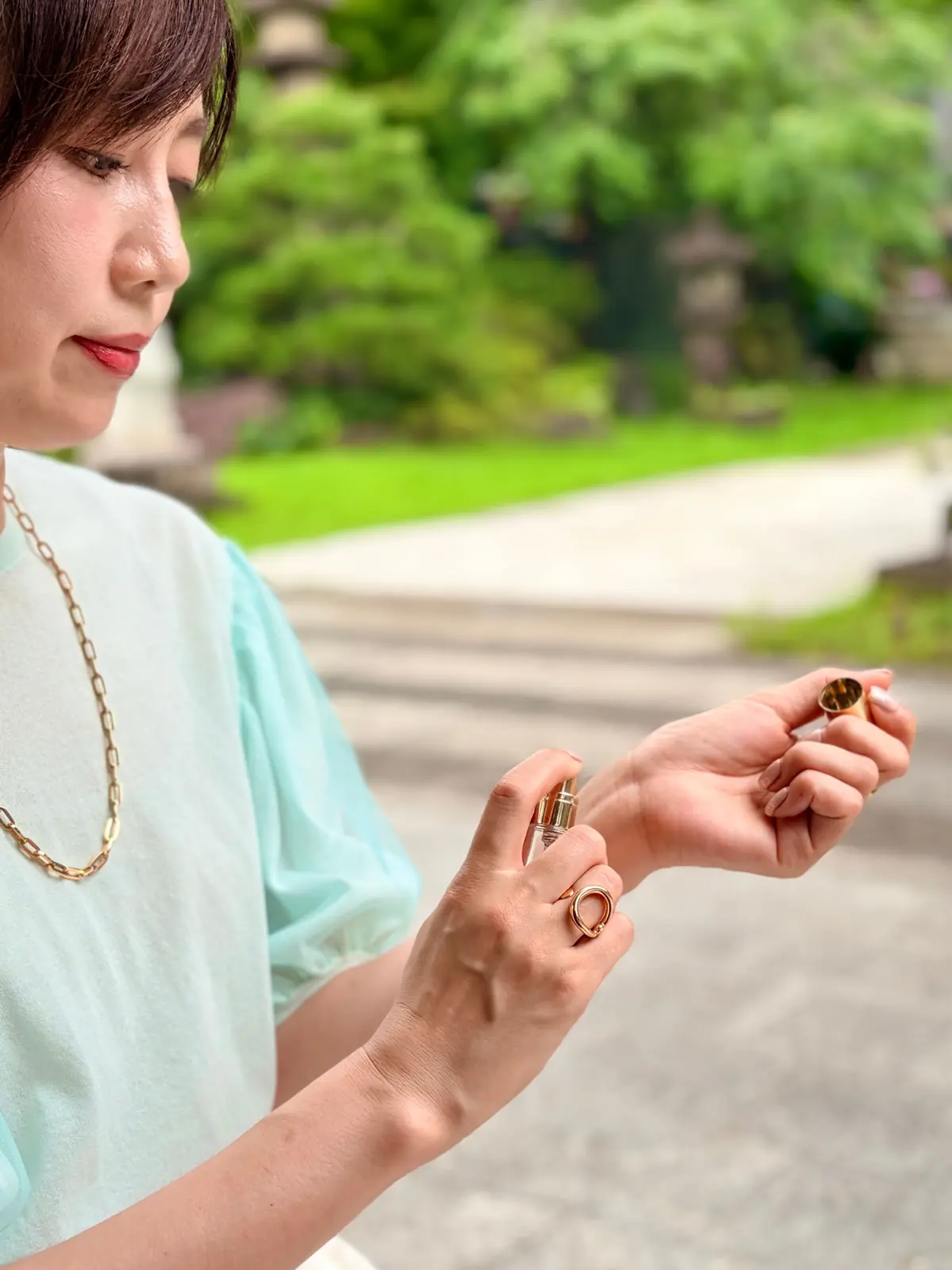 mintグリーンの服を着た女性が、公園の芝生エリアで香水のボトルを手に持ってスプレーしている。