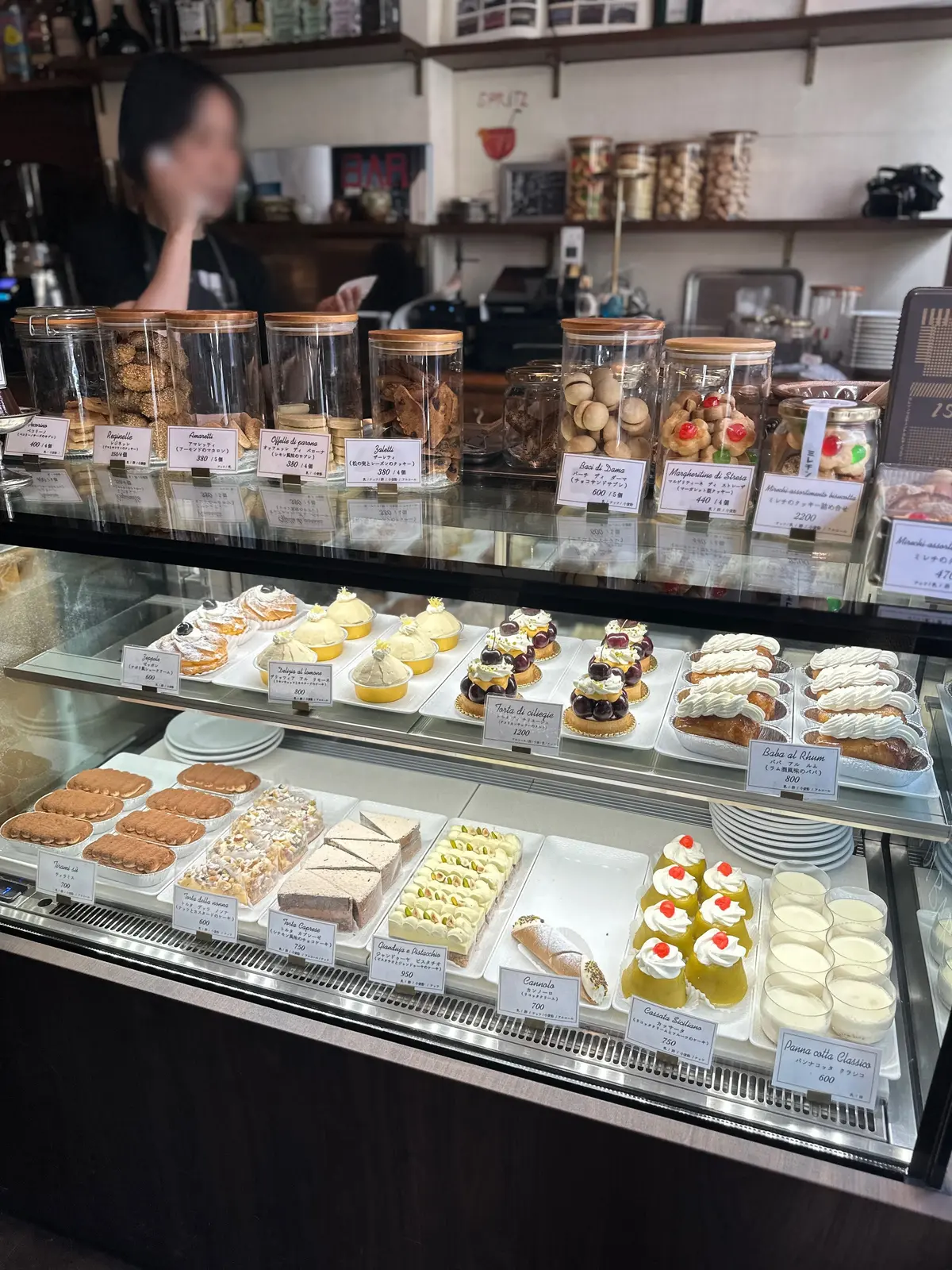 ショーケースに並んだ様々な焼き菓子と瓶に入ったクッキー。タルト、ケーキ、パウンドケーキなど、様々な種類のスイーツが整然と並び、奥には人影が見える。