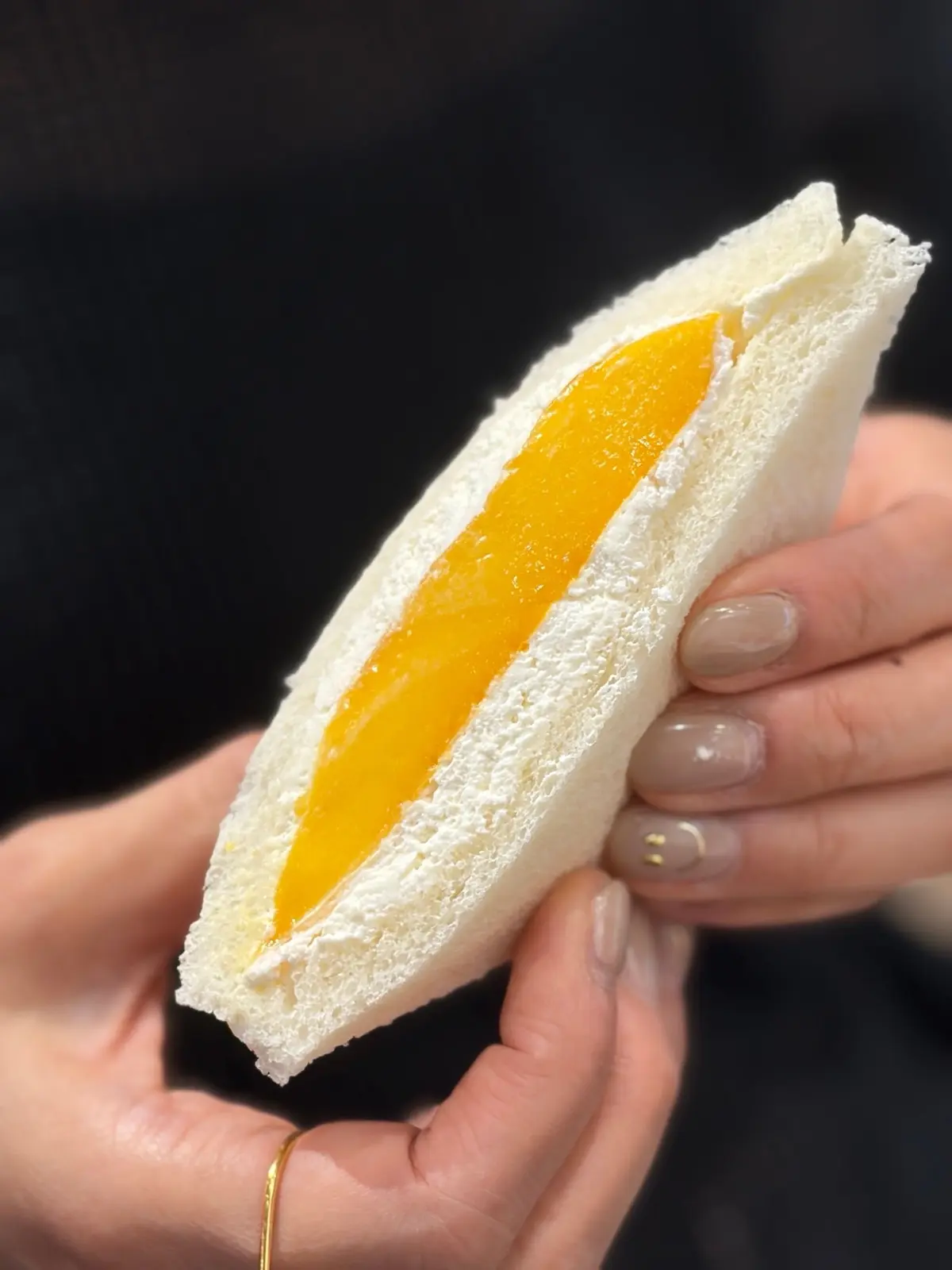手で持ち上げられたサンドイッチ。白い食パンで、中にはオレンジ色の餡が沿うように挟まっている。