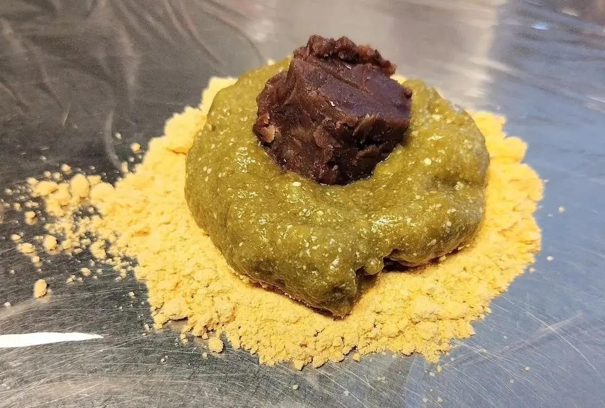緑色の団子に、茶色い餡が乗った和菓子。周りには粉末状の黄色い粉が散らばっている。