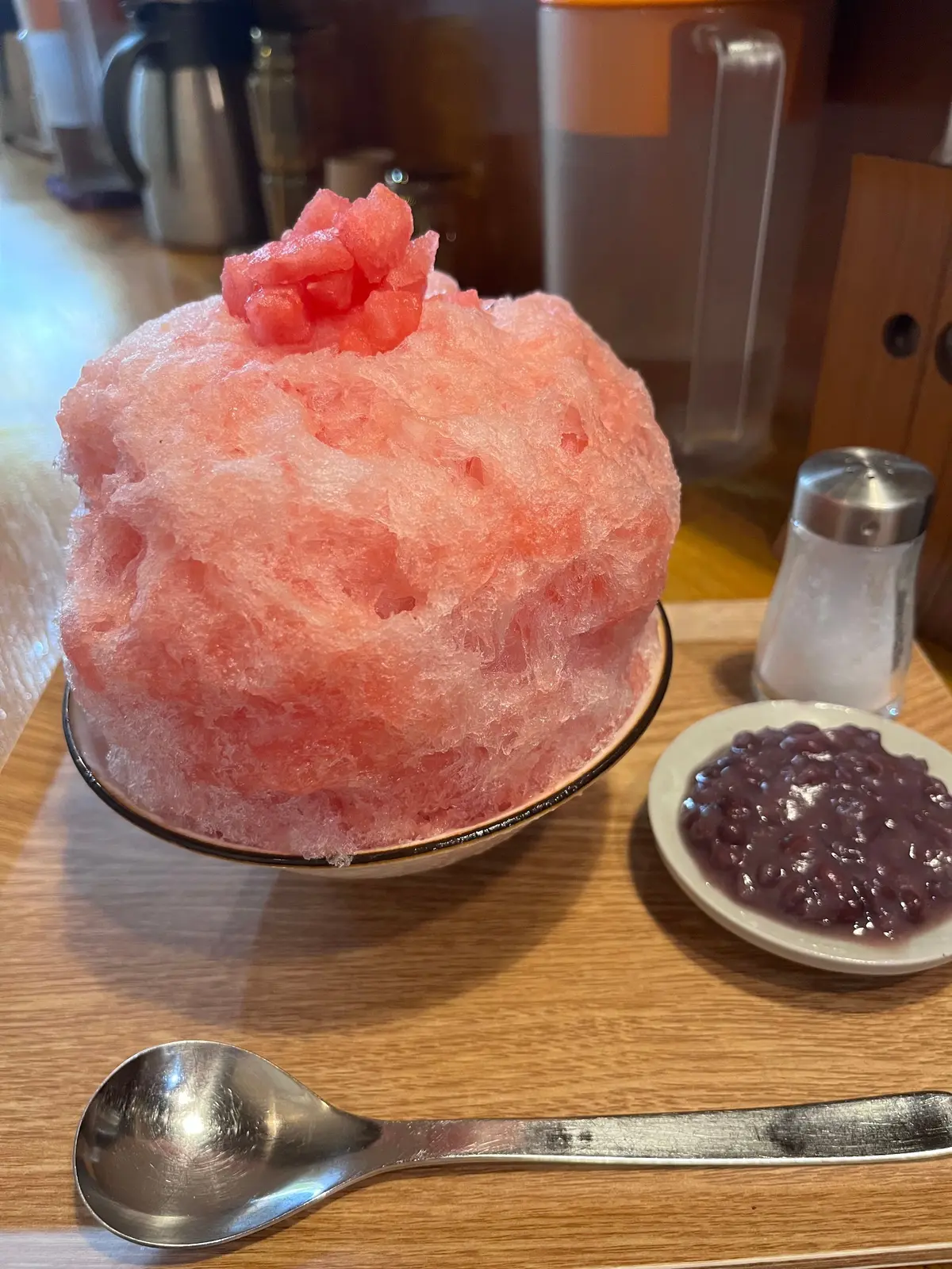 ピンク色のふわふわしたかき氷が黒いお皿に盛り付けられ、右には紫色の豆の小鉢と調味料入れが置かれている。木製のテーブルの上で、銀色のスプーンが横たわっている。