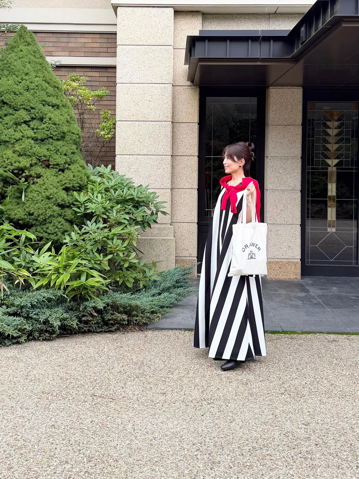 黒と白のストライプのロングドレスを着た女性が、引き戸の前に立っている。赤いスカーフを巻き、白いショッピングバッグを持っている。背景には緑の生垣と砂利敷きの道が見える。