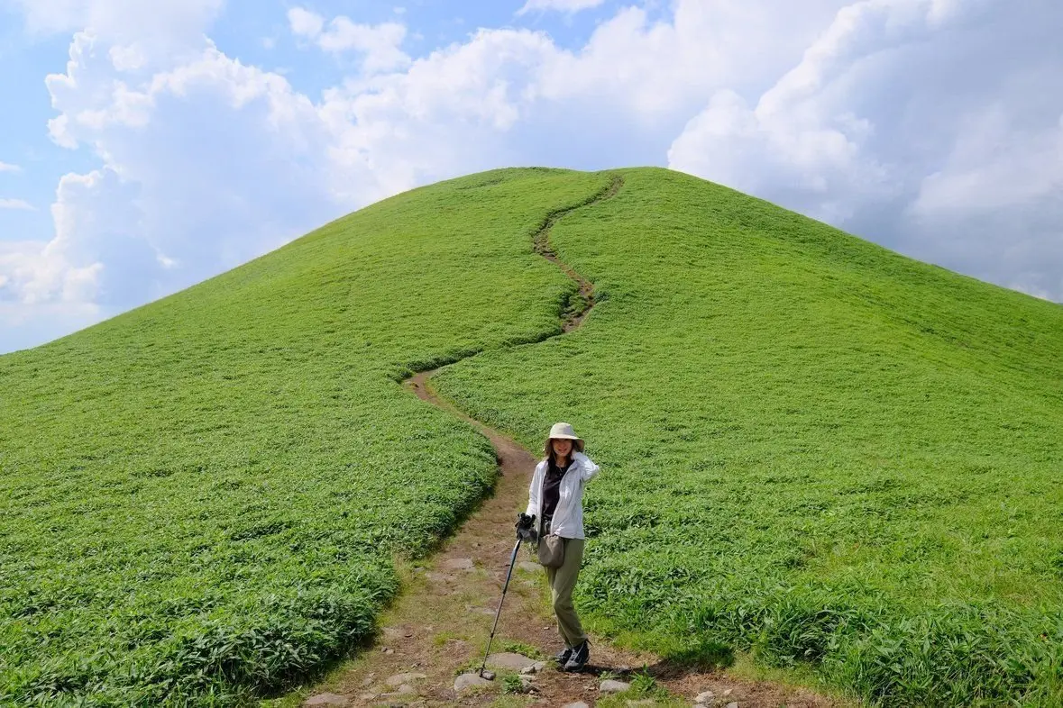 緑の丘に続く道と、帽子をかぶった女性。青空に白い雲が浮かび、晴れた日の風景。