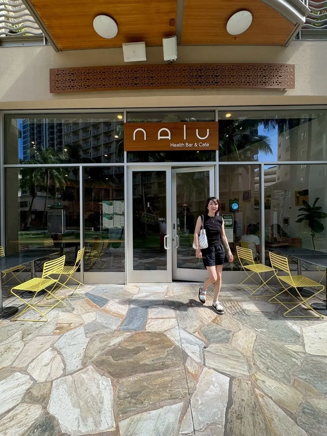 NAIU Health Bar & Caféの入り口前で、石の床の上に黄色い椅子が配置され、店内から明るい光が差し出ている。女性が白いトートバッグを手に歩いている。