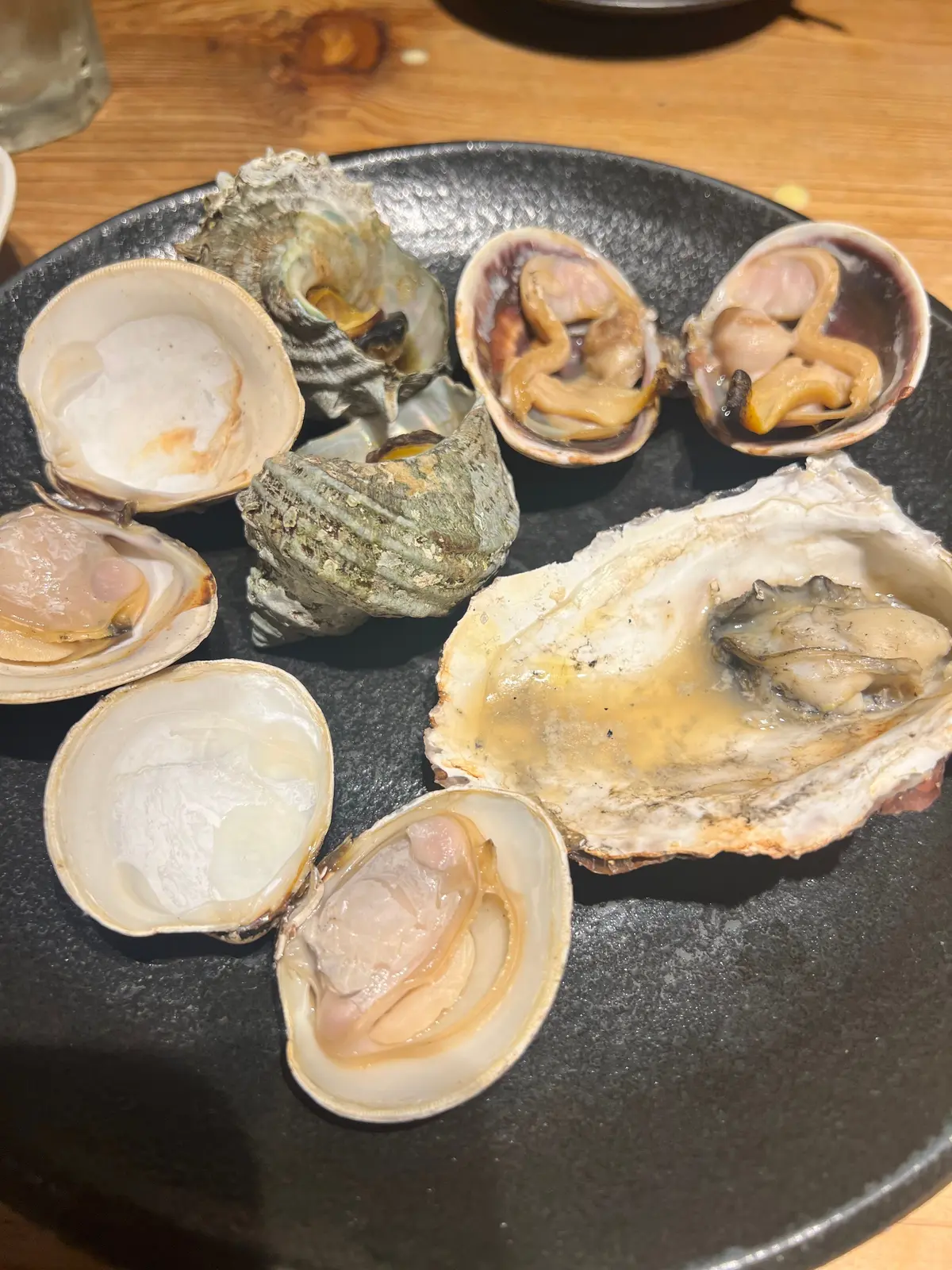 浜焼き。黒いお皿に盛り付けられた、様々な種類の貝。中央にはオイスター、その周りにはアサリやハマグリ、そして緑色の岩ガニのような貝が並んでいる。