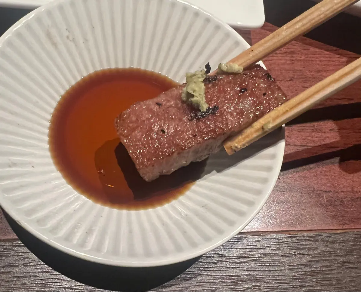 白磁の皿に盛り付けられた、表面に黒胡椒が散らされた赤身の肉の塊。肉の上には緑色のペーストが乗っており、皿には濃い茶色のソースが染み込んでいる。木製の箸で肉が支えられている。