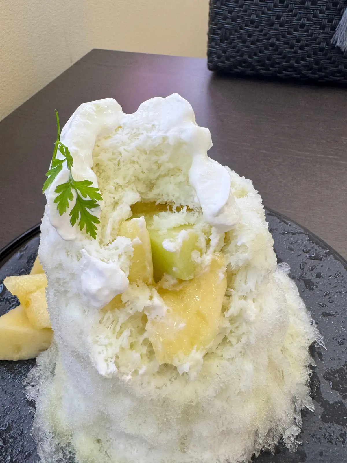 黒いお皿に盛り付けられた、ふわふわの雪氷の上にメロンとクリームが乗ったデザート。メロンの切れ端が横に添えられ、上にはパセリが飾られている。