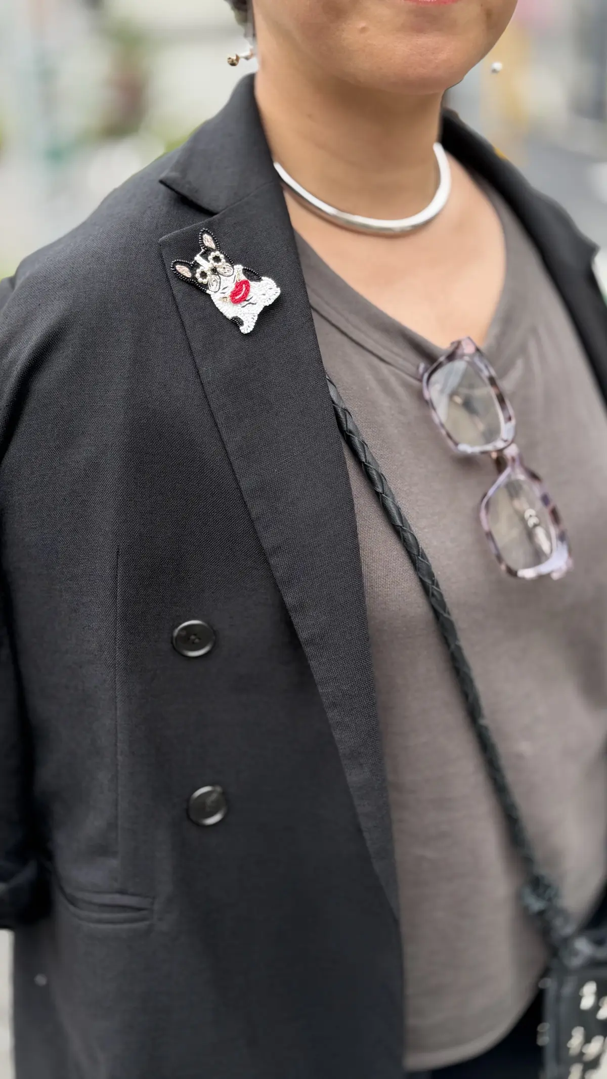 黒いジャケットの襟に、パグの刺繍ブローチが付けられている。グレーのTシャツを着用し、シルバーのチョーカーネックレスを身に着けている。眼鏡をかけた女性のアップ。