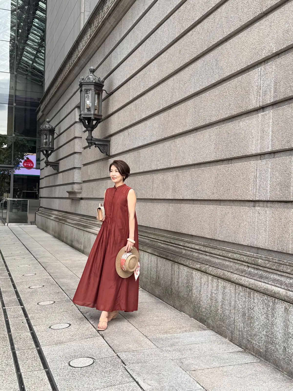 ブラウンのロングワンピースを着た女性が、石造りの建物の前で歩いている。右手に麦わら帽子、左腕にバッグを持つ。背景には現代的な高層ビルが見える。