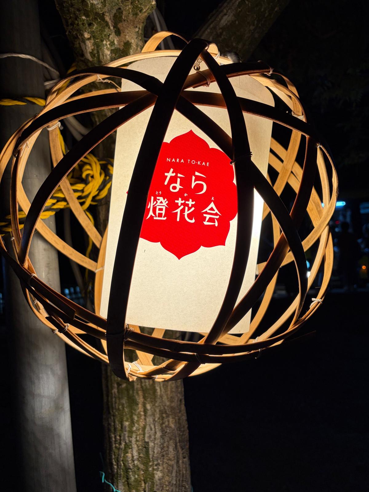 夜の木立に吊るされた、竹製の球体ランタン。中央には赤い円の中に「なら燈花会」と書かれた白い文字が配置されている。球体は複数の竹が交差した構造で、内部は白い紙で覆われている。