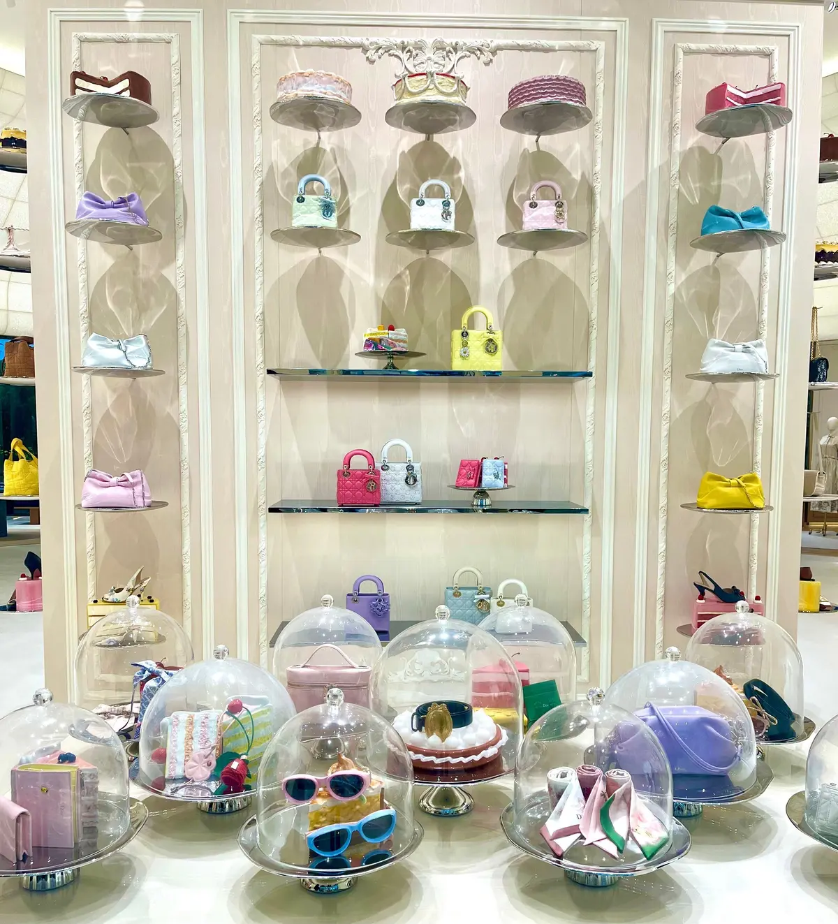 ディオールの展示台と壁面ディスプレイ。ピンクベージュの壁に、様々な色のミニバッグやアクセサリーが棚に並び、ガラスのドームに覆われたケーキに見立てた商品が置かれている。