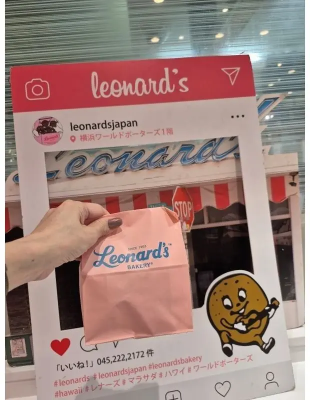 レオンارد'sのピンク色の紙袋を持つ手。背景にはレオンارد'sの店舗の看板と窓が見える。紙袋にはロゴマークが印刷され、右下にはパンのキャラクターが描かれている。
