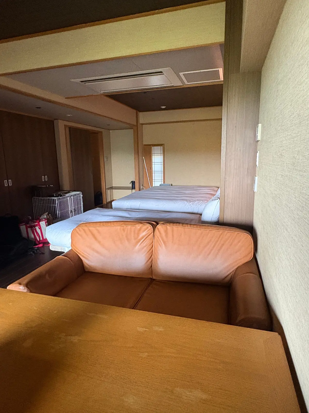 茶色のソファと、その奥に3つのシングルベッドが並ぶ部屋。壁や天井は淡い黄色の木目調。左には収納棚が見える。
