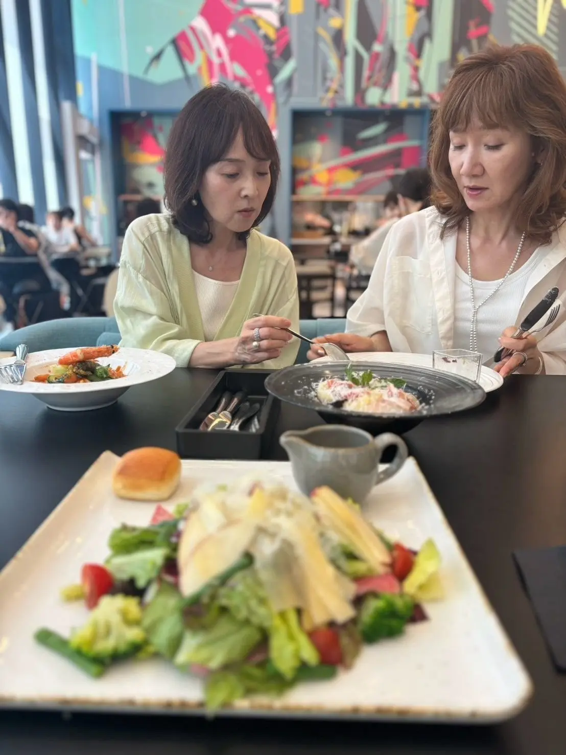 レストランで食事をする2人の女性。左は黄緑色のカーディガン、右は白いシャツを着ている。テーブルにはサラダと料理が並び、背景には抽象的な絵画が飾られている。
