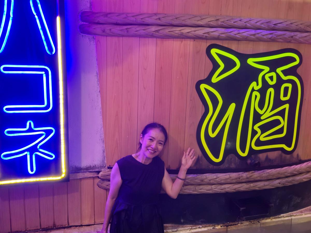 夜のネオンサインと女性。左には青色の「コ」と「ネ」の文字、右には黄色の麺のイラストのネオンサイン。女性は黒い服を着て笑顔で手を振っている。背景は藁で覆われた壁。