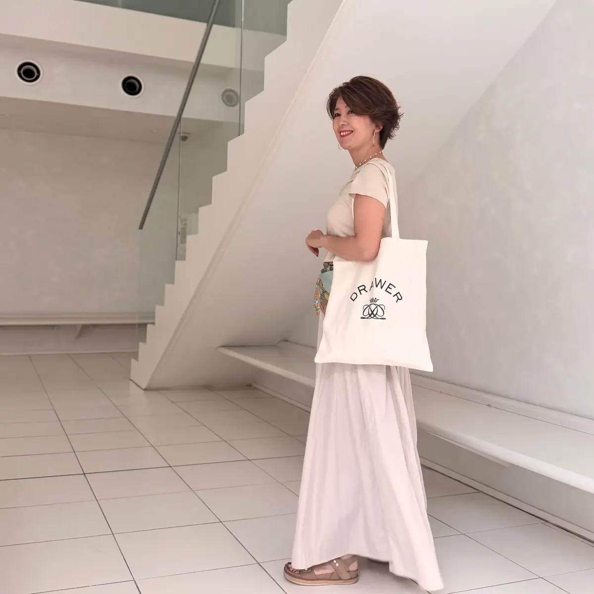 白いワンピースを着た女性が、白い壁と階段の背景で微笑んでいる。左手に白いトートバッグ（DRWERのロゴ入り）を持ち、右手に黄色い袋を持っている。茶色のサンダルを履き、髪は短くカットされている。