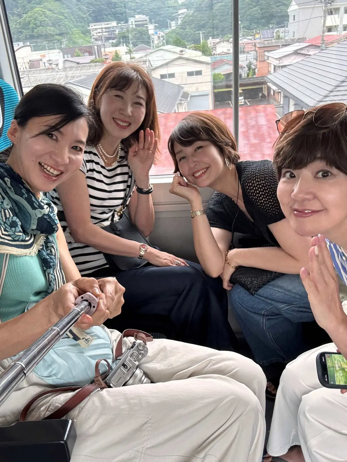 伊豆の踊子号の電車内にて、4人の女性が窓の外の住宅地を見ながら笑う。手前にはカメラと携帯電話が置かれている。