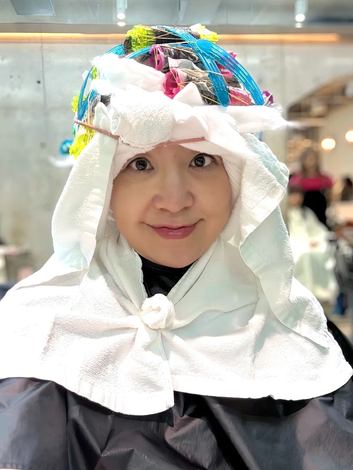 美容院でヘアカラー施術中の女性。白のタオル帽子と黒いビニール製のケープを着用。頭部にはカラフルなヘアカラーシートが覆い被さっている。