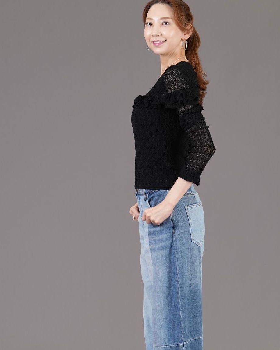黒いレースの長袖トップスと青いデニムスカートを着用した女性が、横を向いて立っている全身写真。茶色の髪を後ろで一つにまとめ、微笑んでいる。背景はグレー。