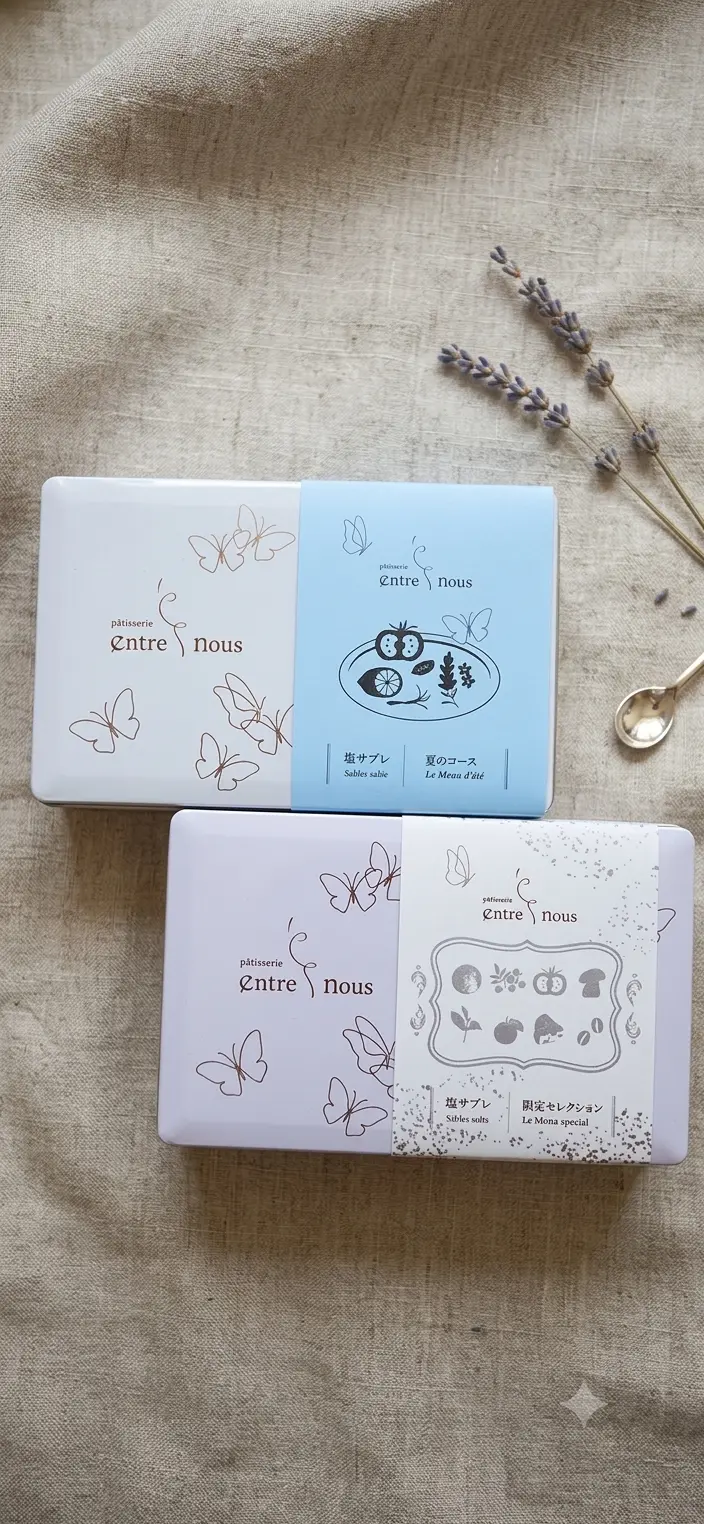 白地の箱に青色の帯が巻かれたお菓子4箱。帯には「patisserie Entre nous」の文字と、それぞれ蝶、フクロウと木の実、蝶のイラストが描かれている。箱の横にはドライフラワーと思われる紫色のものが添えられている。