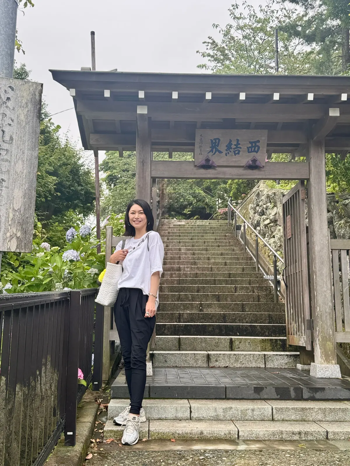女性が石段の最上段に立ち、笑顔でカメラを見つめている。手には白いトートバッグを提げ、白いTシャツと黒いパンツ、ホワイトのスニーカーを着用。手前には木製の鳥居があり、青い看板に「渡部神社」と書かれている。