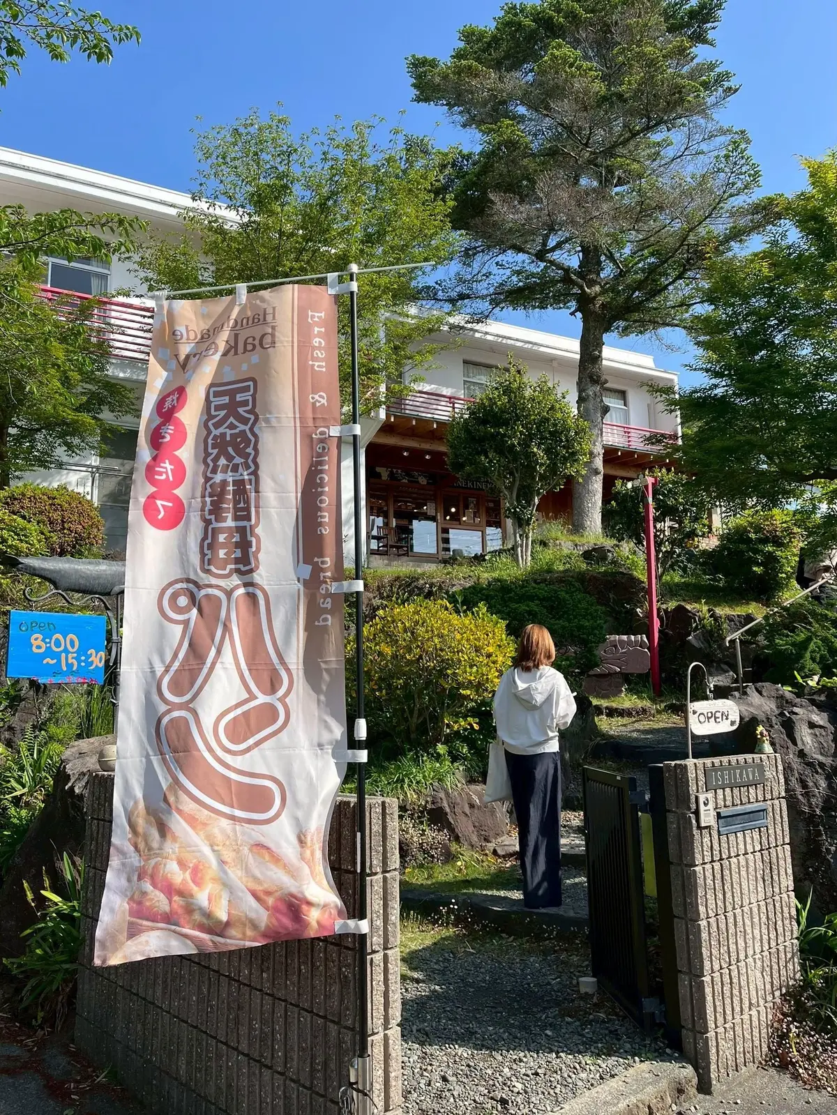 白いフード付きの女性が、緑豊かな庭園を通り、石造りの門をくぐり、レストランの入り口へ向かう。左側に「天空のラーメン」の横断幕が設置され、開店時間表示がある。