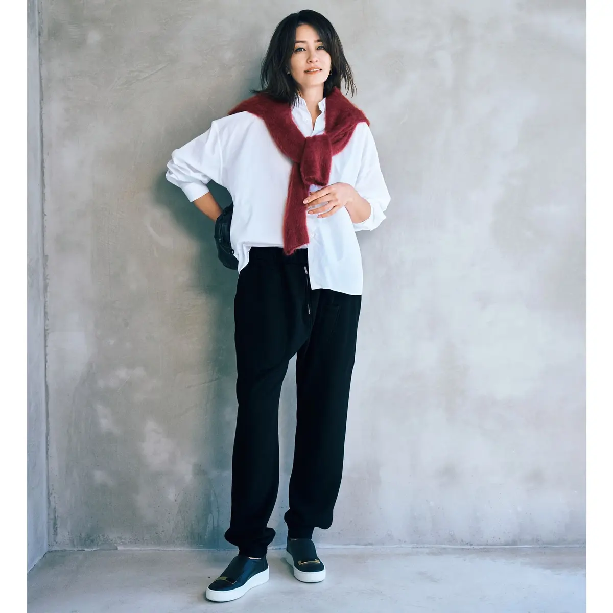 白シャツに黒のスウェットパンツ、赤いふわふわのショールを巻いた女性。黒いスリッポンを履き、背景はグレーのコンクリート壁。