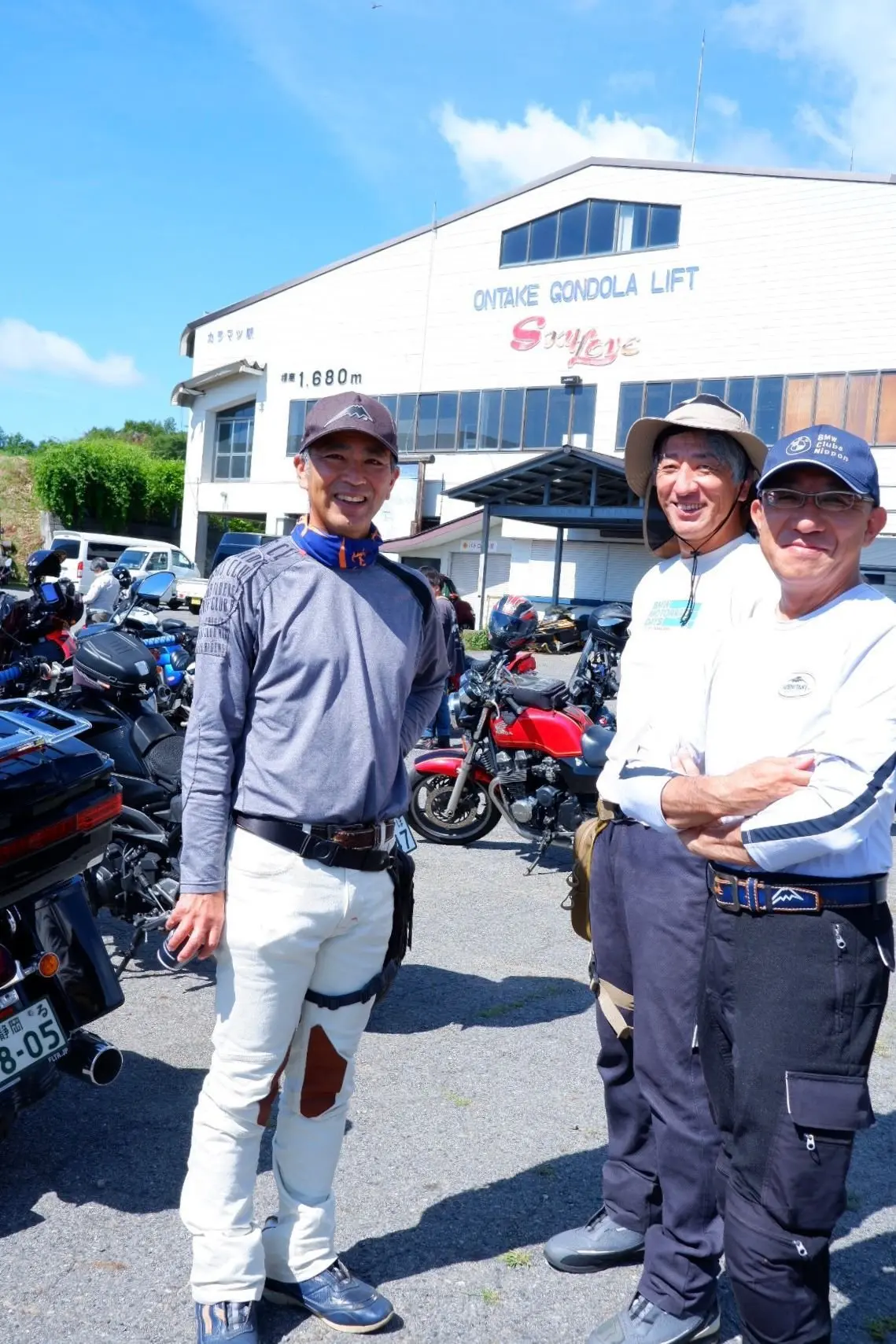 晴天の下、バイクのグループが建物の前で記念撮影をしている。左から若い女性、男性2人。背景には複数のバイクが並び、白い建物には「ONTAKE GONDOLA LIFT Skyeye」の文字が見える。
