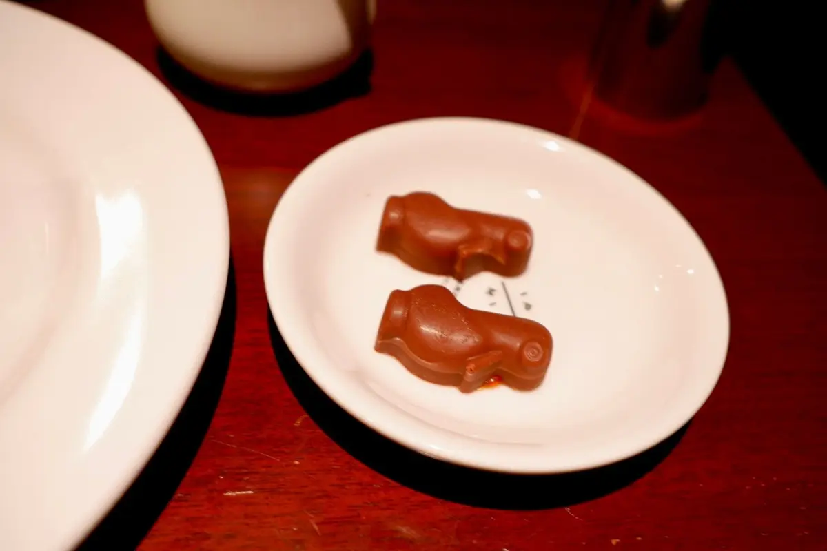 赤いチョコレート菓子が2つ、白い楕円形の皿に置かれている。背景には、別の皿と茶色の飲み物が写っている。
